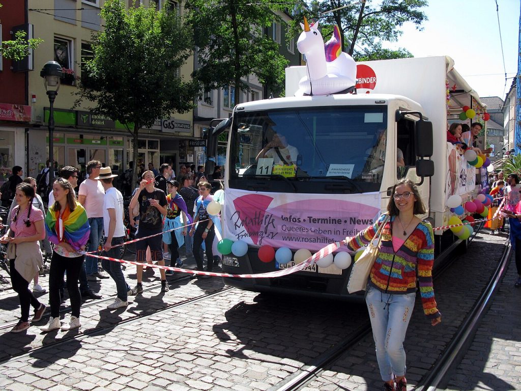 Ein mit bunten Luftballons geschmückter Lastwagen fährt durch die Freiburger Innenstadt umgeben von Teilnehmenden der CSD Parade, vorne Angebracht ist das Logo der Rosa Hilfe