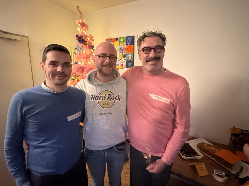 Drei Männer stehen nebeneinander, einer trägt einen blauen Pullover, einer einen weißen Hard-Rock-Café-Hoodie, einer einen rosa Pullover, im Hintergrund ein geschmückter rosa Weihnachtsbaum und bunte Wandbilder