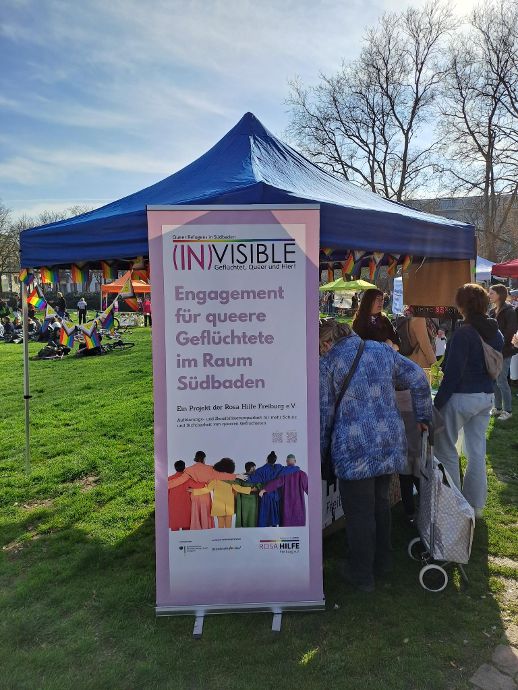Foto eines Pavillons (Infostand), an dem verschiedene Personen stehen (von hinten abgebildet). In der Bildmitte zu sehen ist die Beach Flag des Projekts Invisible mit Projektlogo und mehreren sich umarmenden Personen verschiedener Ethnien, die in Regenbogenfarben gekleidet sind.