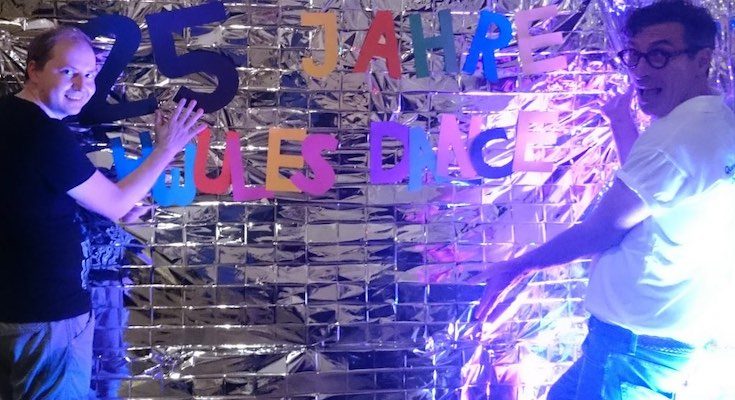 Zwei Personen stehend lachend vor einer silber verhängten Wand, auf der in bunten Buchstaben 25 Jahre Schwulesdance steht.