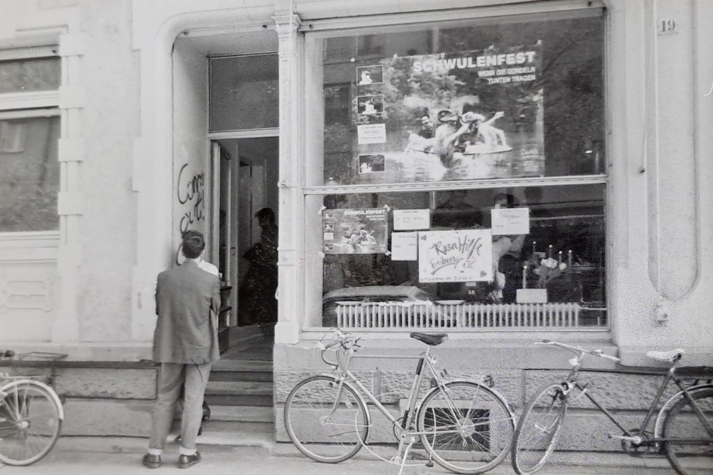 Schwarz-weiß Foto der Außenseite der neuen Räume in der Eschholzstraße, Am Fenster angebracht ist ein Schild mit der Aufschrift ,,Rosa Hilfe Freiburg e.V.", darüber Plakat der Veranstaltung ,,Wenn die Gondeln Tunten tragen"