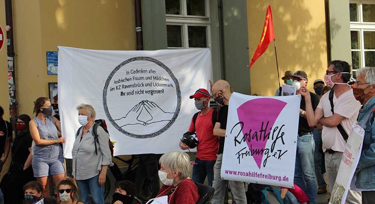 Personen mit Masken auf der Straße mit großen Bannern, eins davon mit Rosa Hilfe Logo