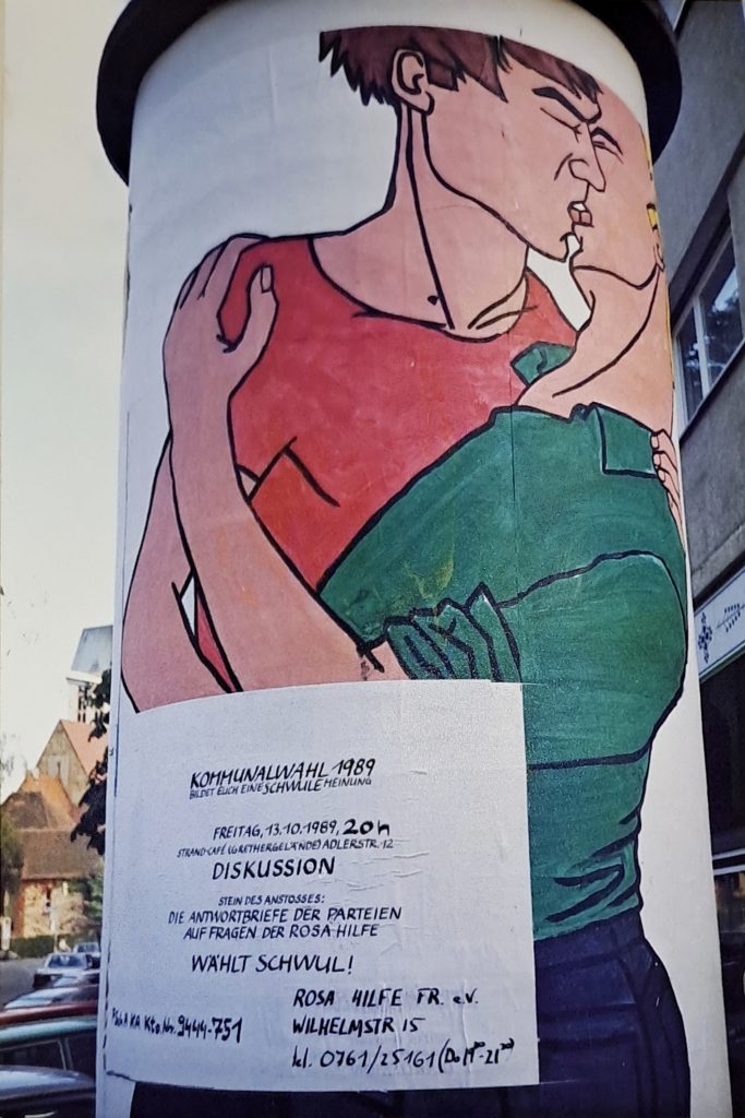 Litfasssäule mit zwei sich küssenden gezeichneten Männern, Poster zur Veranstaltung ist sichtbar.