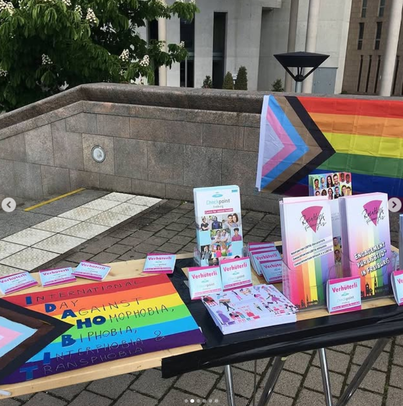 Foto von Infostand mit Flyern von Checkpoint und Rosa Hilfe, im Hintergrund ist eine große Progress pride Fahne aufgehängt