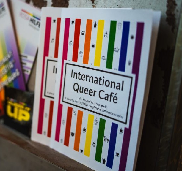Foto des Flyers des international queer Cafe (weiß mit regenbogenbunten Streifen und illustrierten Teekannen) in einem Flyerregal)