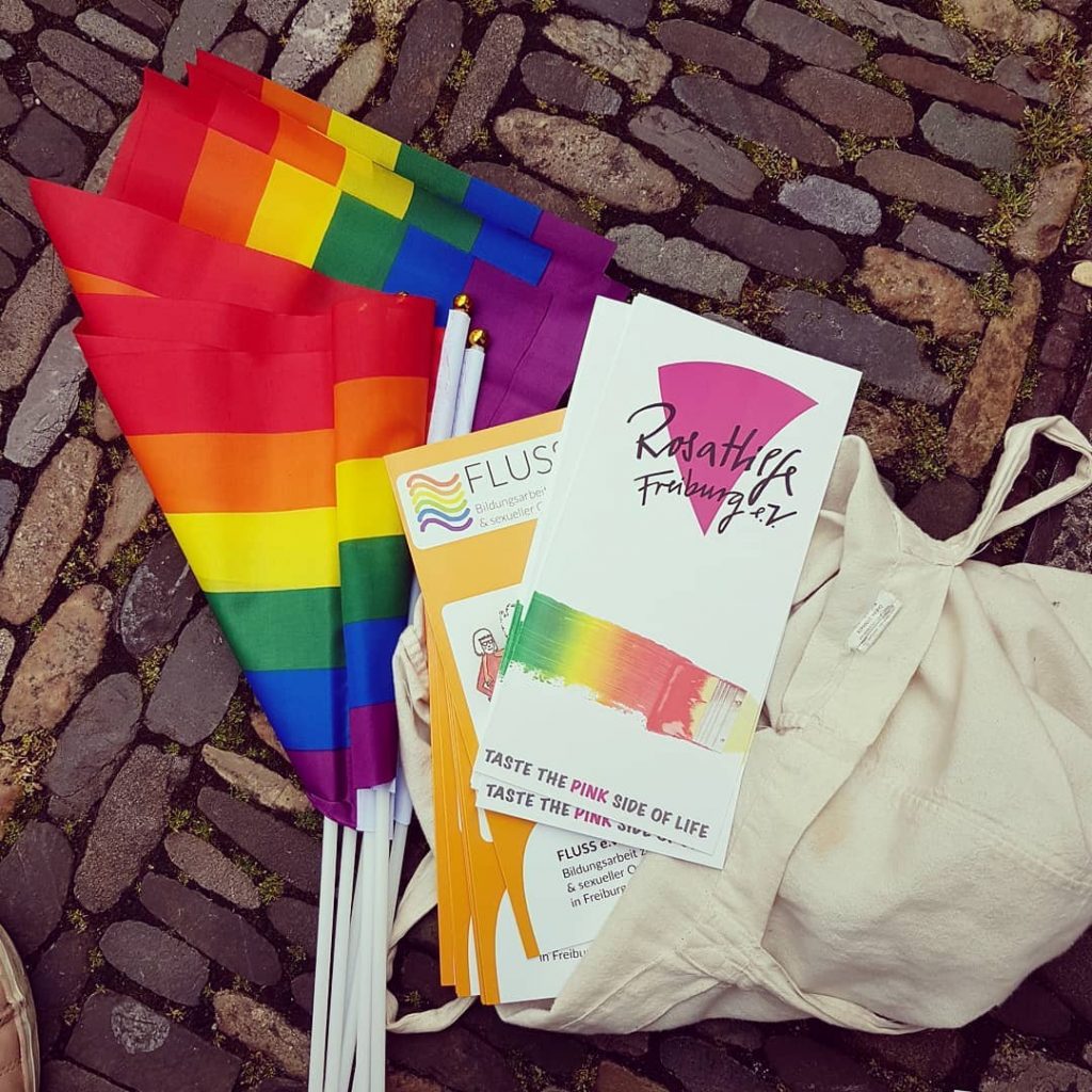 Auf Kopfsteinpflaster liegen mehrere kleine Regenbogenflaggen und Flyern von Fluss und Rosa Hilfe auf einem weißen beutel.