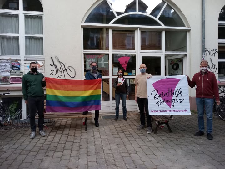 Fünf Personen mit medizinischer Maske stehen vor der Fassade der Rosa Hilfe mit Logo-Banner und einer Regenbogenfahne.