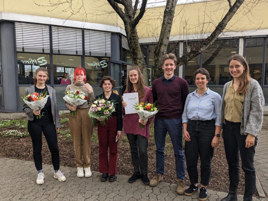 Sieben Personen stehen nebeneinander in einer Reihe und lächeln in die Kamera, die vier auf der linken Seite halten jeweils einen Blumenstrauß und eine zusätzlich eine Urkunde in der Hand.