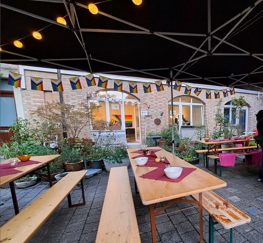 Einige Bierbänke stehen unter einem blauen Pavillon, der mit Lichterketten und einer Girlande aus Progress Pride Fahnen geschmückt ist.